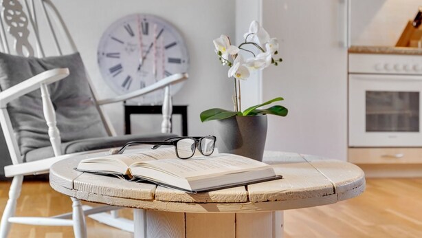 Ein runder Holztisch mit offenem Buch und Brillen, einer Topf-Orchidee und einer Uhr an der Wand.