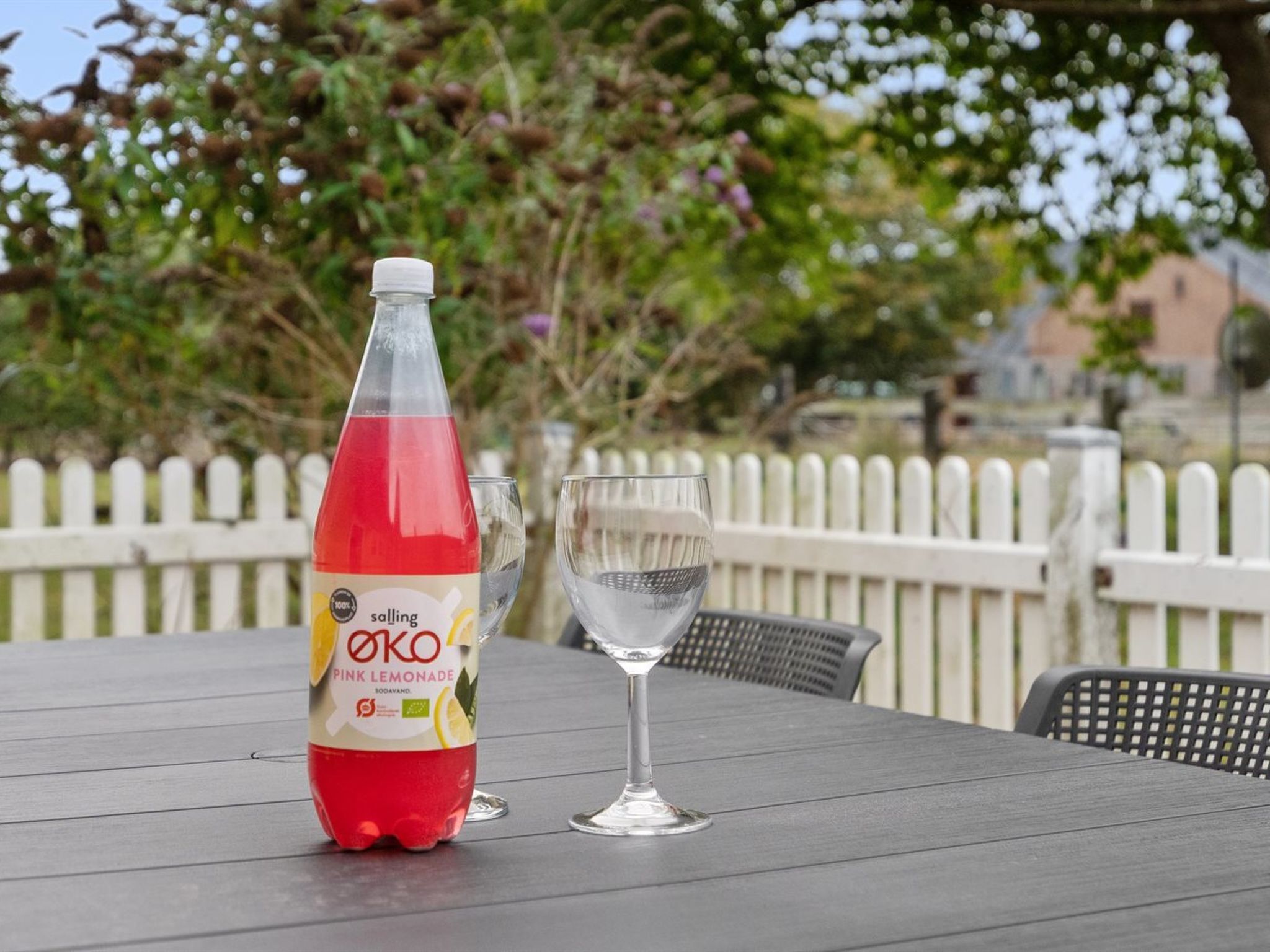 A bottle of pink lemonade and a glass on an outdoor table with a white picket fence in the background.