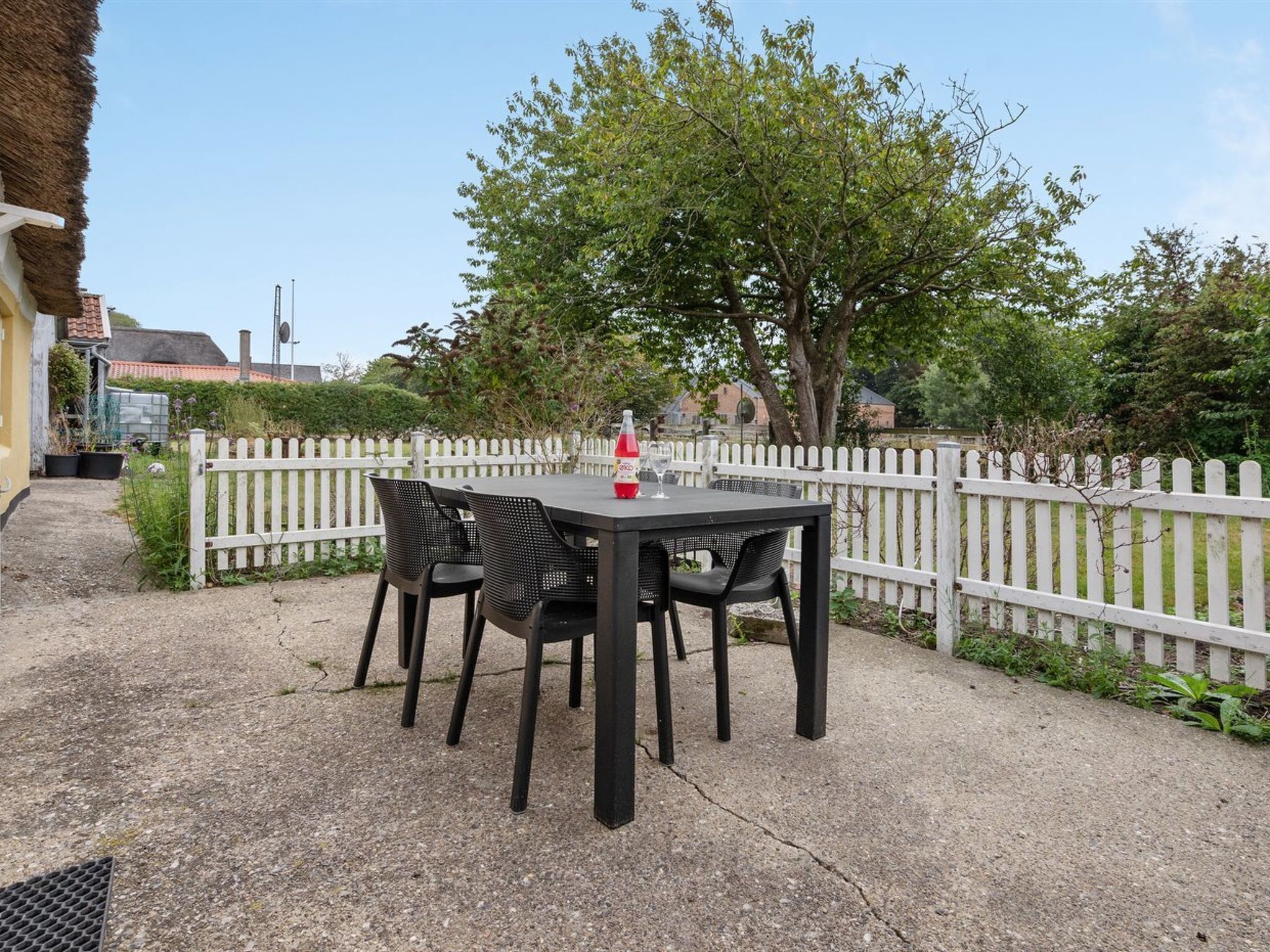 Un patio al aire libre con una mesa y cuatro sillas, una botella de bebida roja, y una cerca blanca de piquetes al fondo.