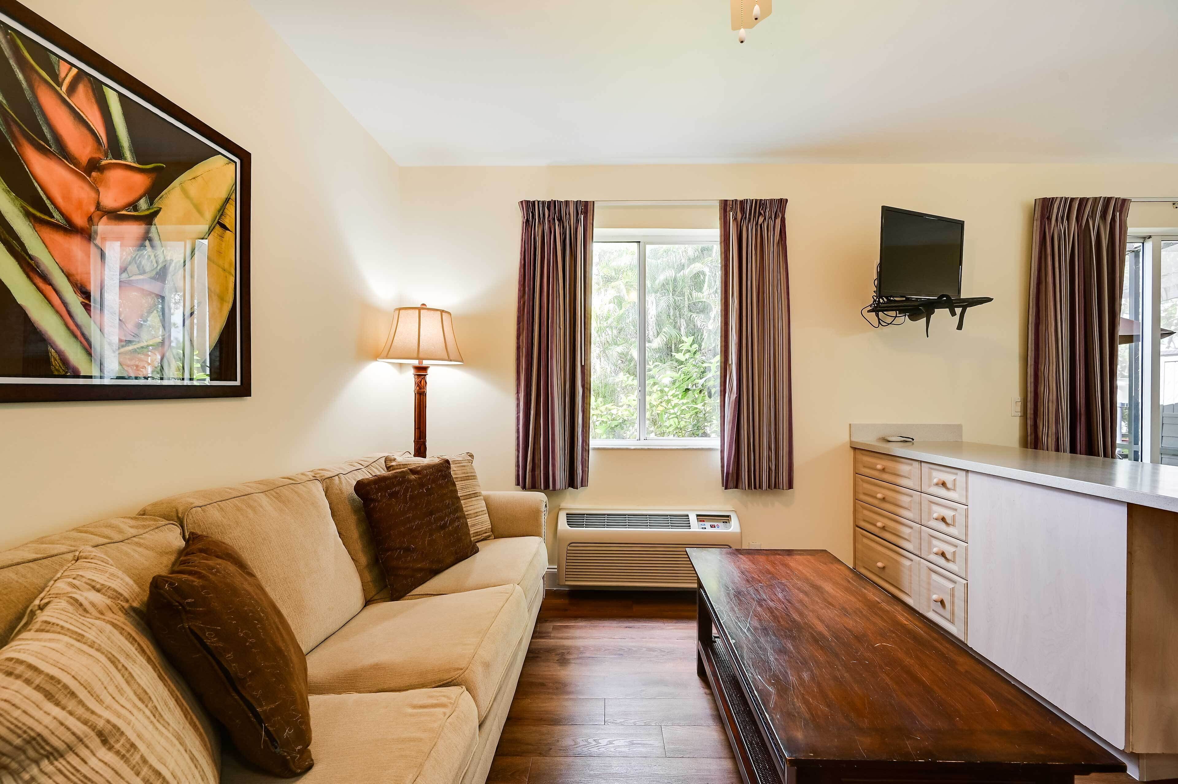 Ein Zimmer mit einem beige farbenen Sofa, einem Holztisch, einem an der Wand montierten Flachbildfernseher, einem Fenster mit Vorhängen und einer kleinen Küchenzeile mit Arbeitsfläche und Schubladen.