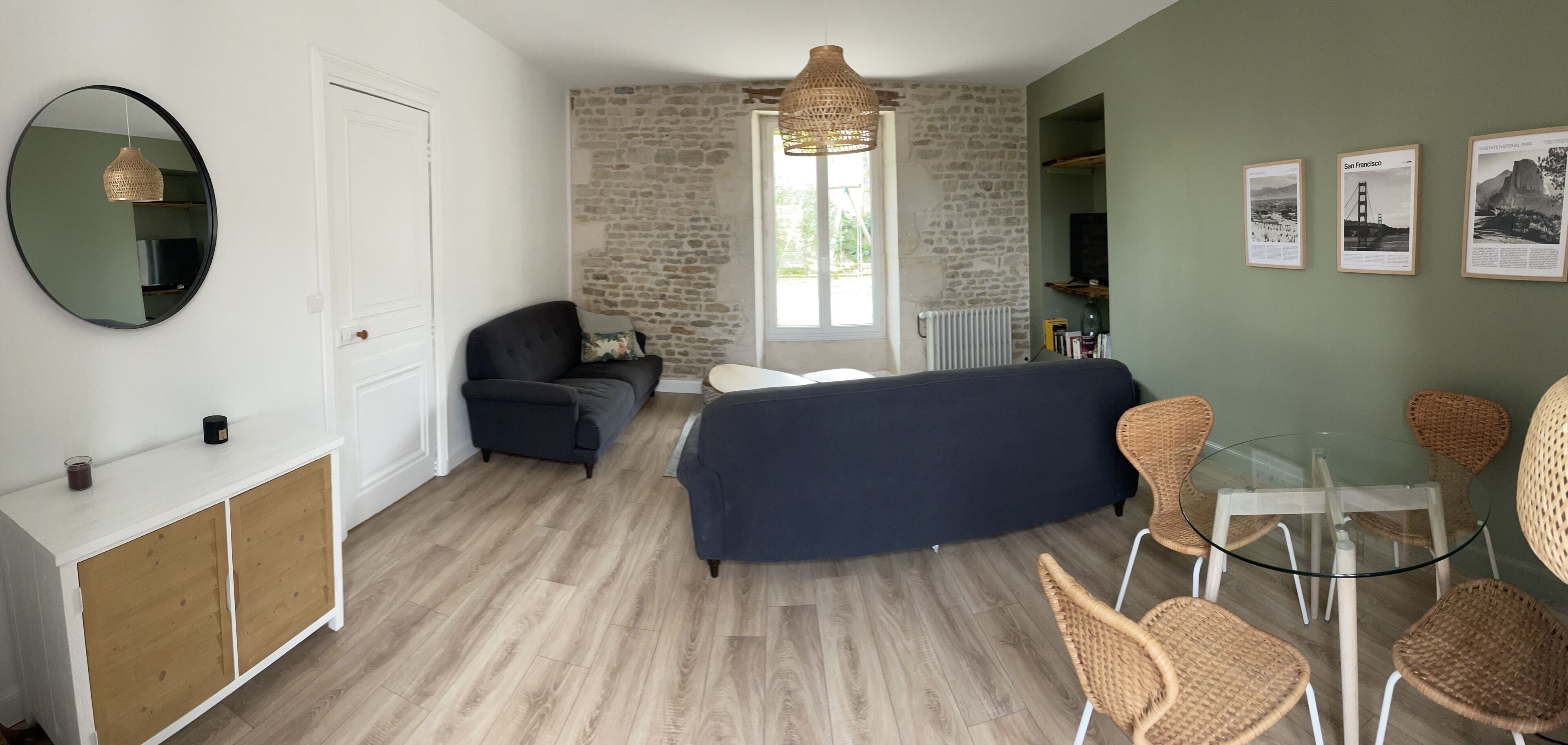 A room with a stone wall, a bed, a sofa, a glass table with chairs, a round mirror, and a white door.