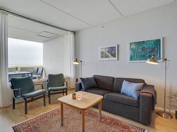 A modern living room with a grey sofa, two armchairs, a wooden coffee table, and a patterned rug. There are two floor lamps and framed pictures on the wall.