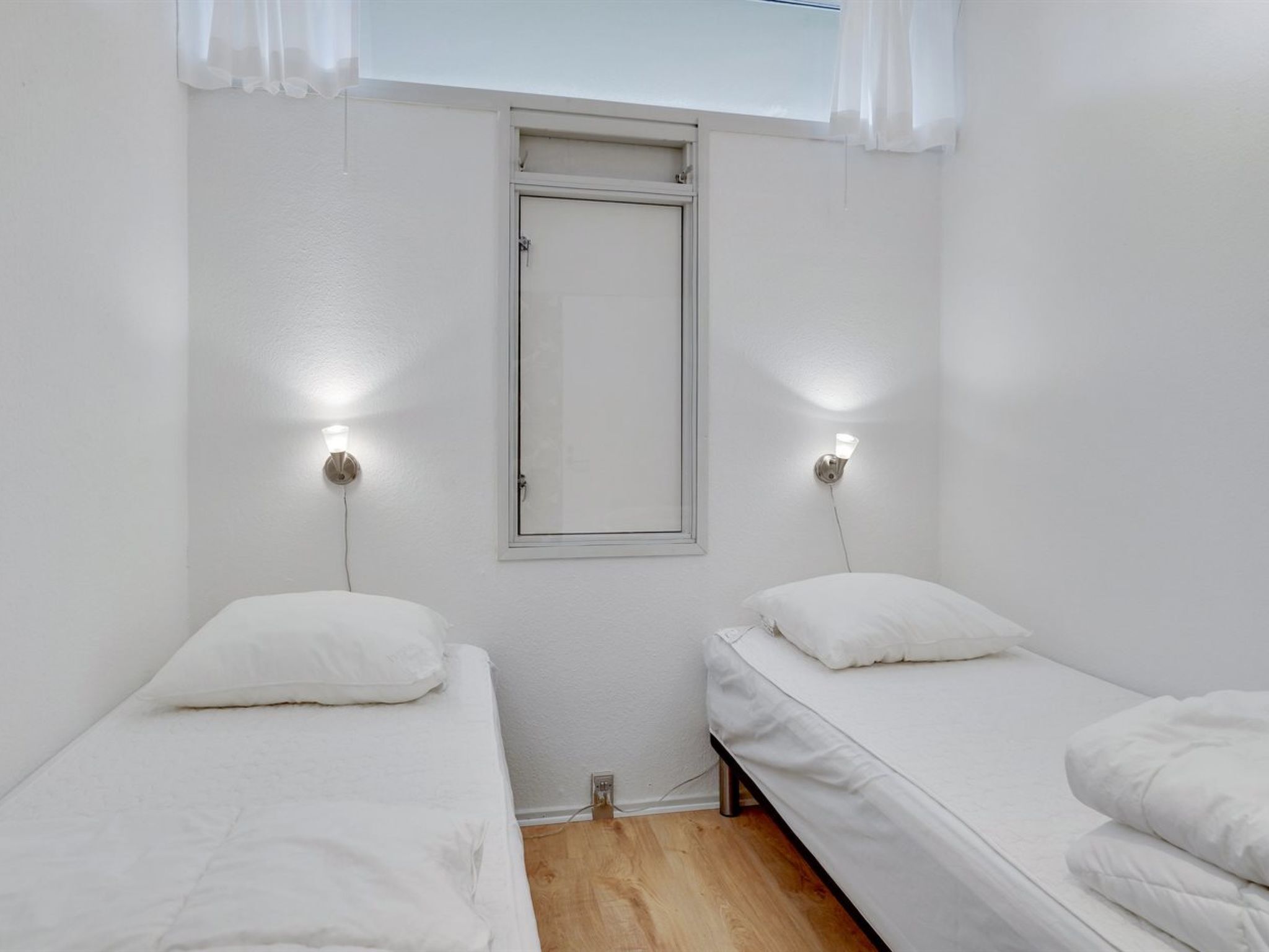 A small, clean room with two single beds, a window with a white frame, and wall-mounted lights.