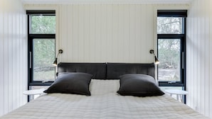 Ein Bett mit weißer, gesteppter Bettdecke, zwei schwarzen Kissen und einem dunklen Kopfteil. Zwei Nachttische mit Lampen stehen bereit, und der Raum hat weiße Wände und große Fenster.