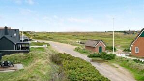 A rural road with houses on either side, a grassy field, and a small thatched-roof building.