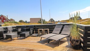 Une terrasse en bois, aménagée avec du mobilier d’extérieur, notamment des chaises longues et une table, qui offre une vue sur un quartier résidentiel avec ses maisons et un ciel dégagé.