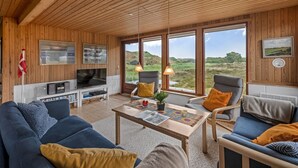 Ein gemütliches Wohnzimmer mit Fernseher, Sofa und Sesseln, große Fenster mit Blick auf die Landschaft.