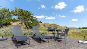 Eine Holzterrasse mit Gartenmöbeln, darunter Stühle und ein Tisch, eingebettet in eine natürliche Landschaft unter blauem Himmel.