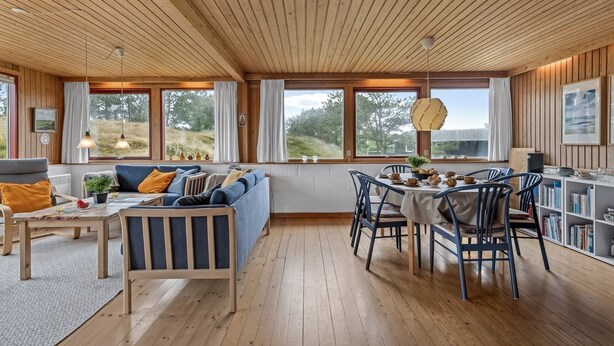 Ein geräumiger Wohnbereich mit Holzboden, einer Sofagarnitur, einem Esstisch mit Stühlen und großen Fenstern mit Blick ins Freie.