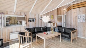 A cozy living room with a black leather sofa, wooden coffee table, and a chair. A window with a view of trees is visible, and the room has wooden walls and a sloped ceiling. - "Germaine" - 1.2km from the sea by Interhome (Fanø)