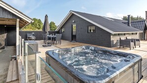 A modern outdoor hot tub area with a wooden deck, a dining set, and a small building with a dark roof.
