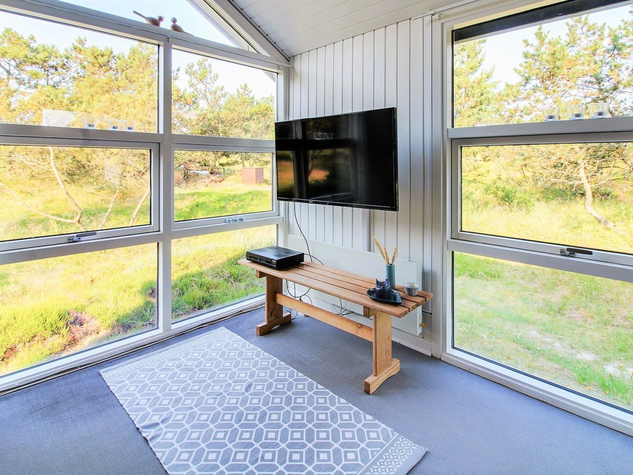 A room with large windows, a flat-screen TV mounted on the wall, a wooden bench, and a patterned rug.