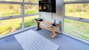 Una habitación con ventanas amplias, un televisor de pantalla plana en la pared, un banco de madera y un tapete estampado.