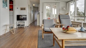 A modern living room with a wooden floor, a TV, two grey armchairs, a dining table with fruit, and large windows.