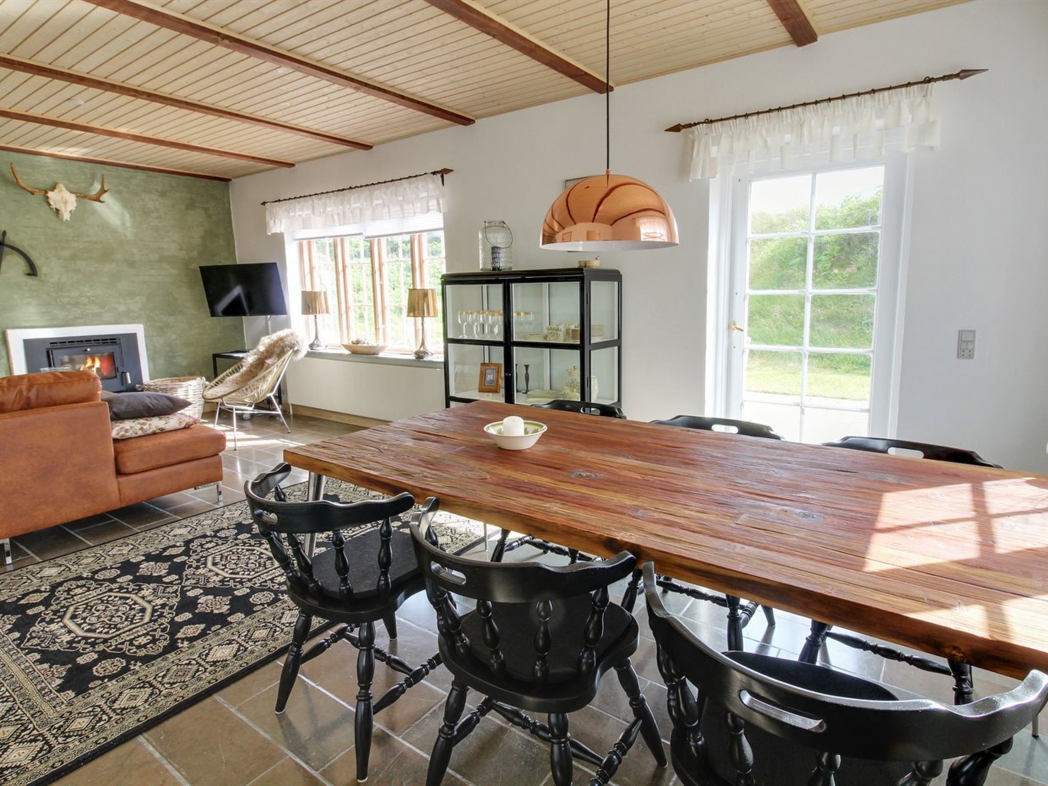 A spacious living room with a large wooden dining table, black chairs, a fireplace, a television, and a glass cabinet.