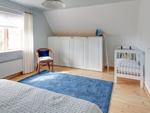 Un dormitorio con techo inclinado, suelo de madera, cama con colcha acolchada, alfombra azul, silla de madera, cuna blanca y armario.