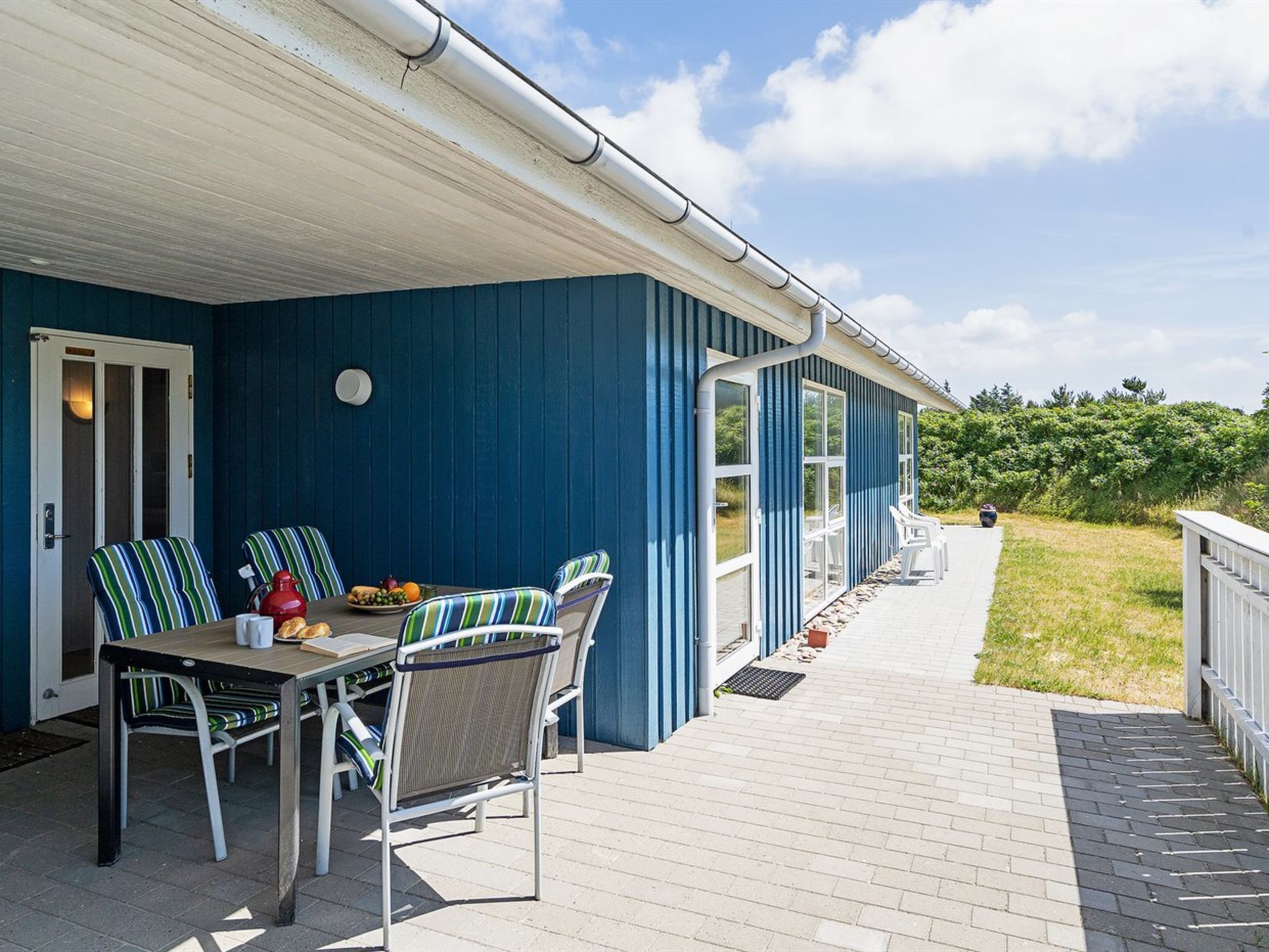 A covered patio with a table and chairs, a door, and a pathway leading to a deck.