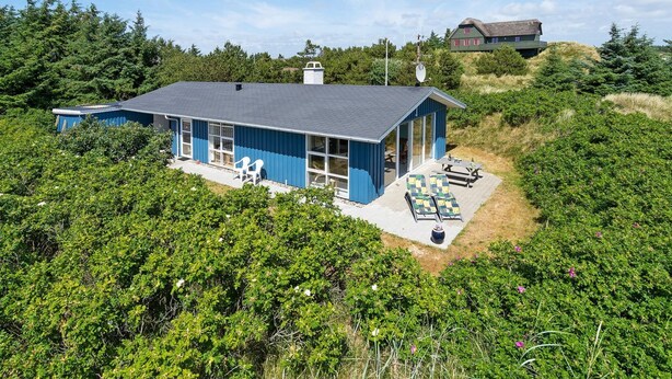 Une maison bleue dotée d’une grande terrasse et d’un espace salon extérieur, le tout entouré d’une végétation luxuriante.