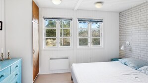 Une chambre à coucher comprenant un lit, une commode, deux fenêtres avec des rideaux, un radiateur et une lampe fixée au mur.