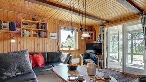 Un salón acogedor con techo de madera, un sofá de cuero negro, una mesa de centro con tazas y platos, una estantería y un televisor.