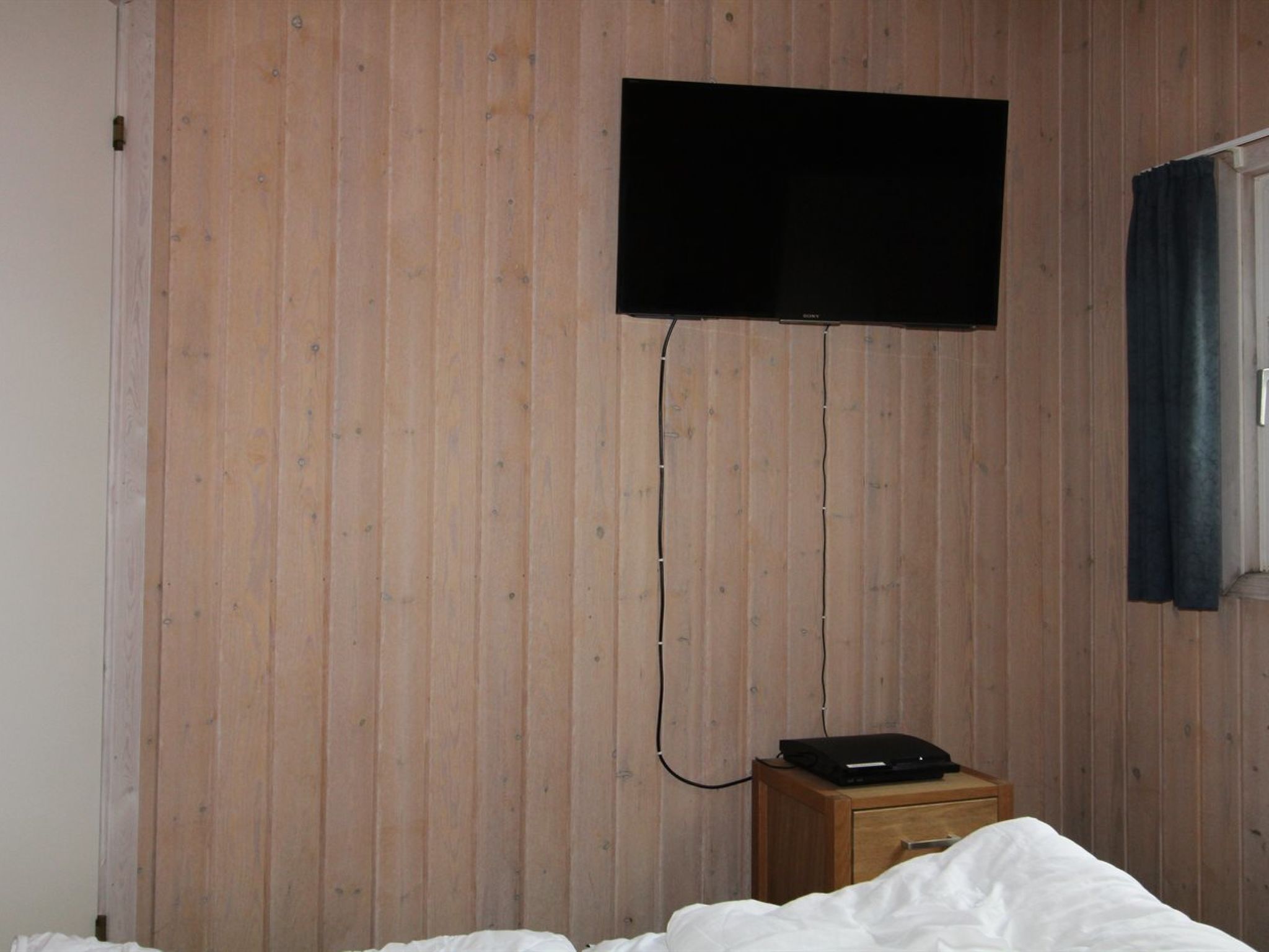 Et værelse med trævæg, et fladskærms-tv ophængt på væggen, et trænatbord med en bærbar computer og en seng med hvide sengetøj.