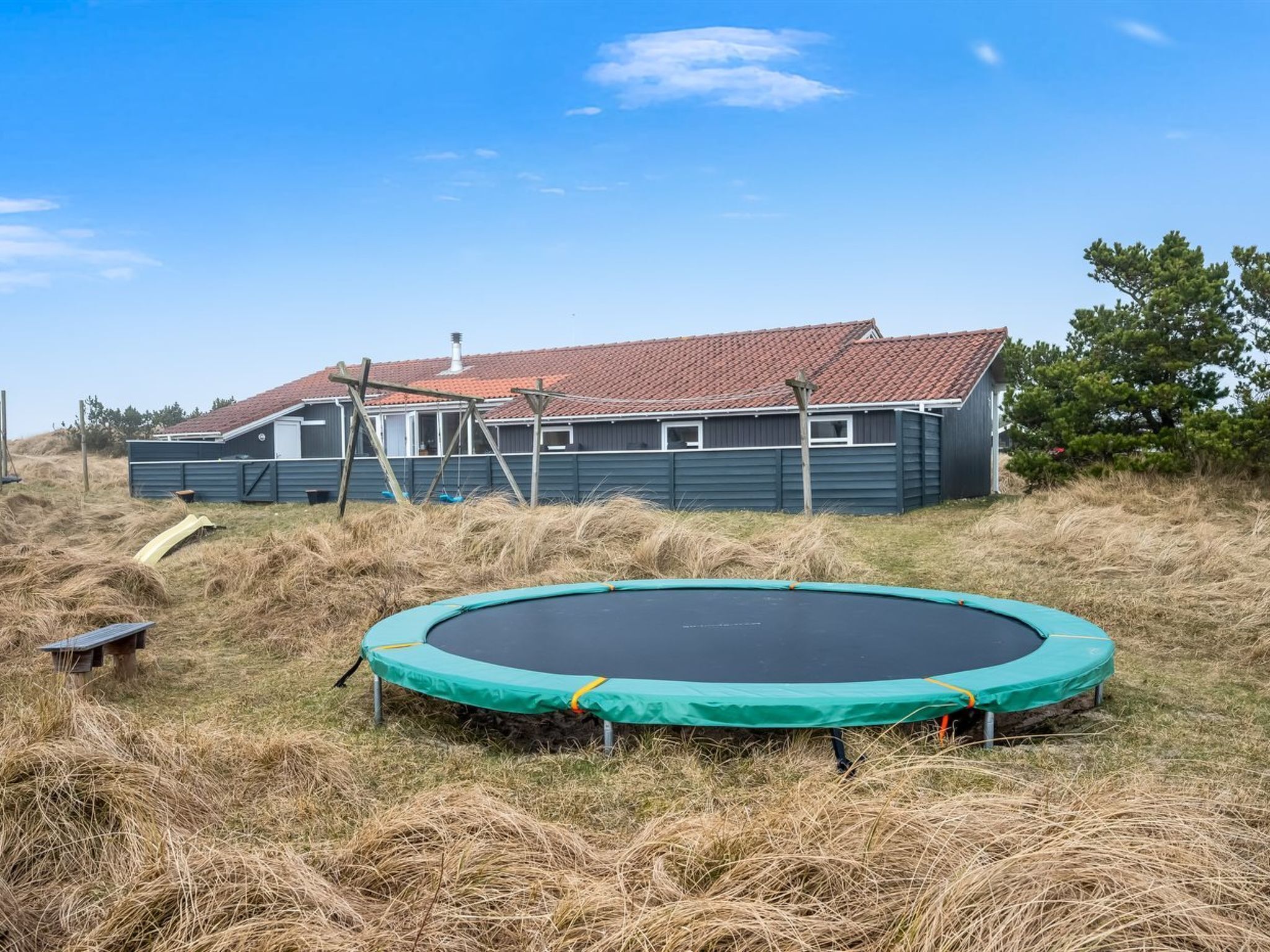 Ein Haus mit rotem Ziegeldach, ein Trampolin im Vordergrund und eine hölzerne Bank.