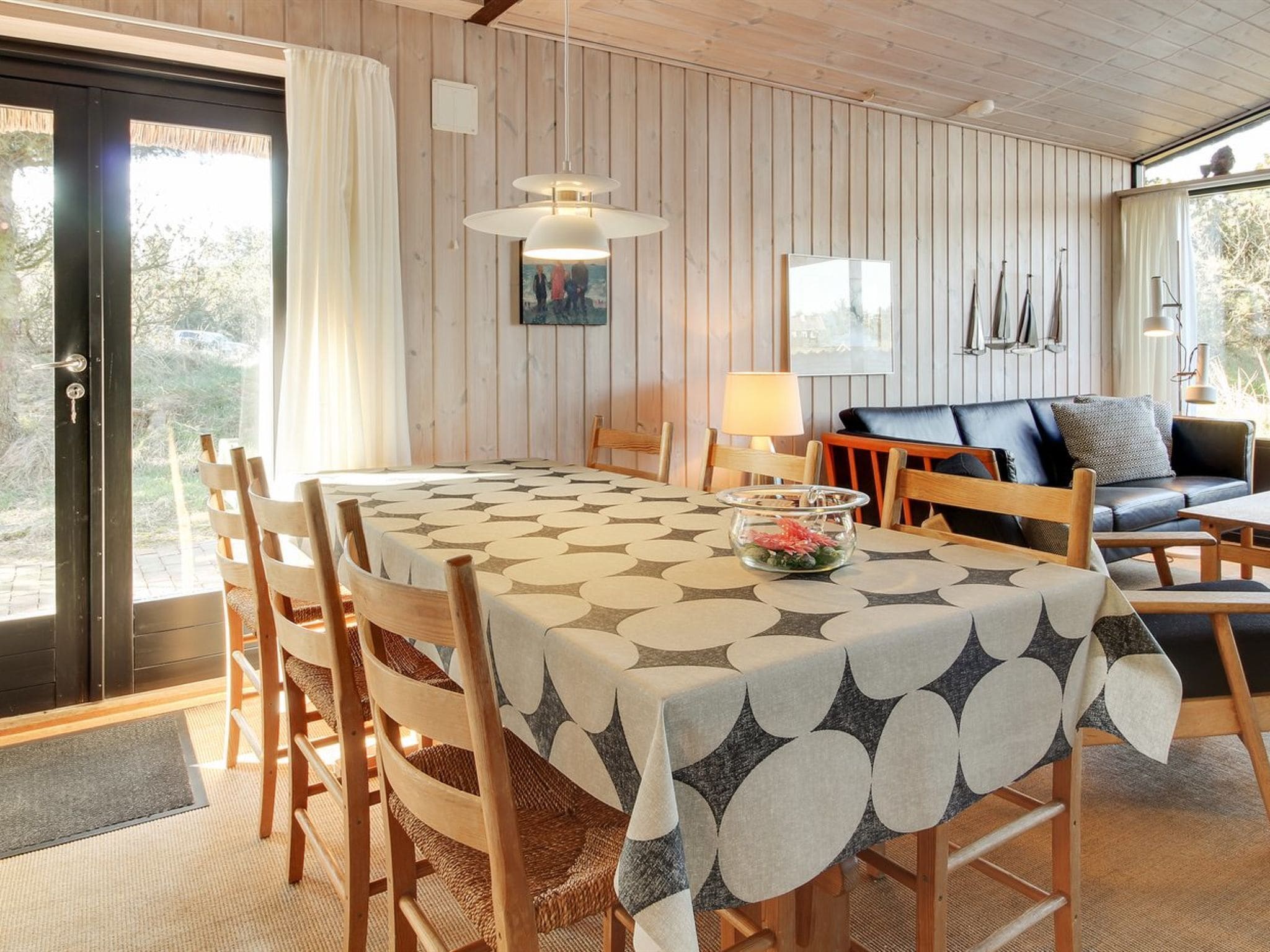 Ein Essbereich mit einem Tisch, bedeckt mit einer gemusterten Tischdecke, Holzstühle, eine schwarze Couch mit Kissen und eine Pendelleuchte.