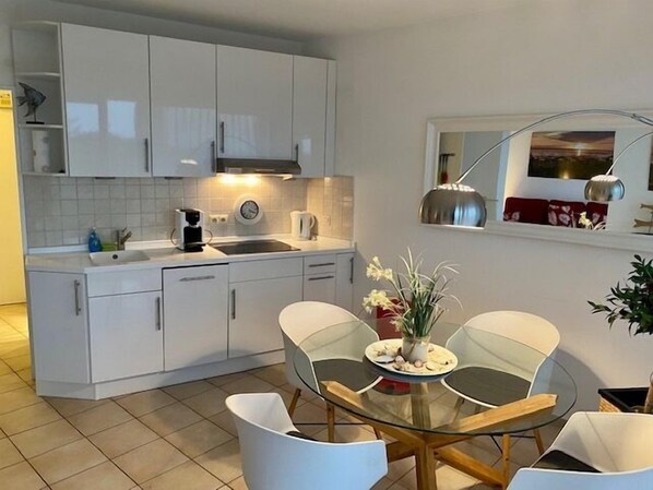 A modern kitchen with white cabinets, a sink, and a coffee maker. There is a dining table with chairs and a vase of flowers. - Seehütte Ferienwohnung 702 (Cuxhaven)