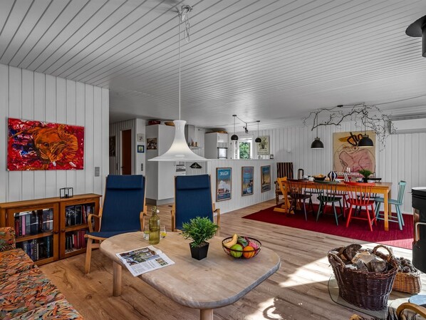 A spacious living room with a wooden floor, a dining area with a red rug, and a kitchen in the background.