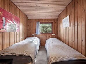 Eine Holzhütte mit zwei Einzelbetten, einem Fenster mit Vorhang und einem gerahmten Bild an der Wand.