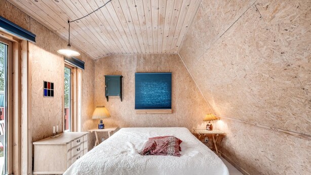 Un dormitorio con techo de madera, una cama con ropa de cama blanca, una mesita de noche con l ámpara y una ventana con vistas a árboles.