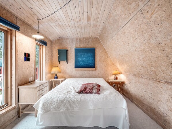 Ein Schlafzimmer mit Holzboden, einem Bett mit weißer Bettwäsche, einem Nachttisch mit Lampe und einem Fenster mit Blick auf Bäume.
