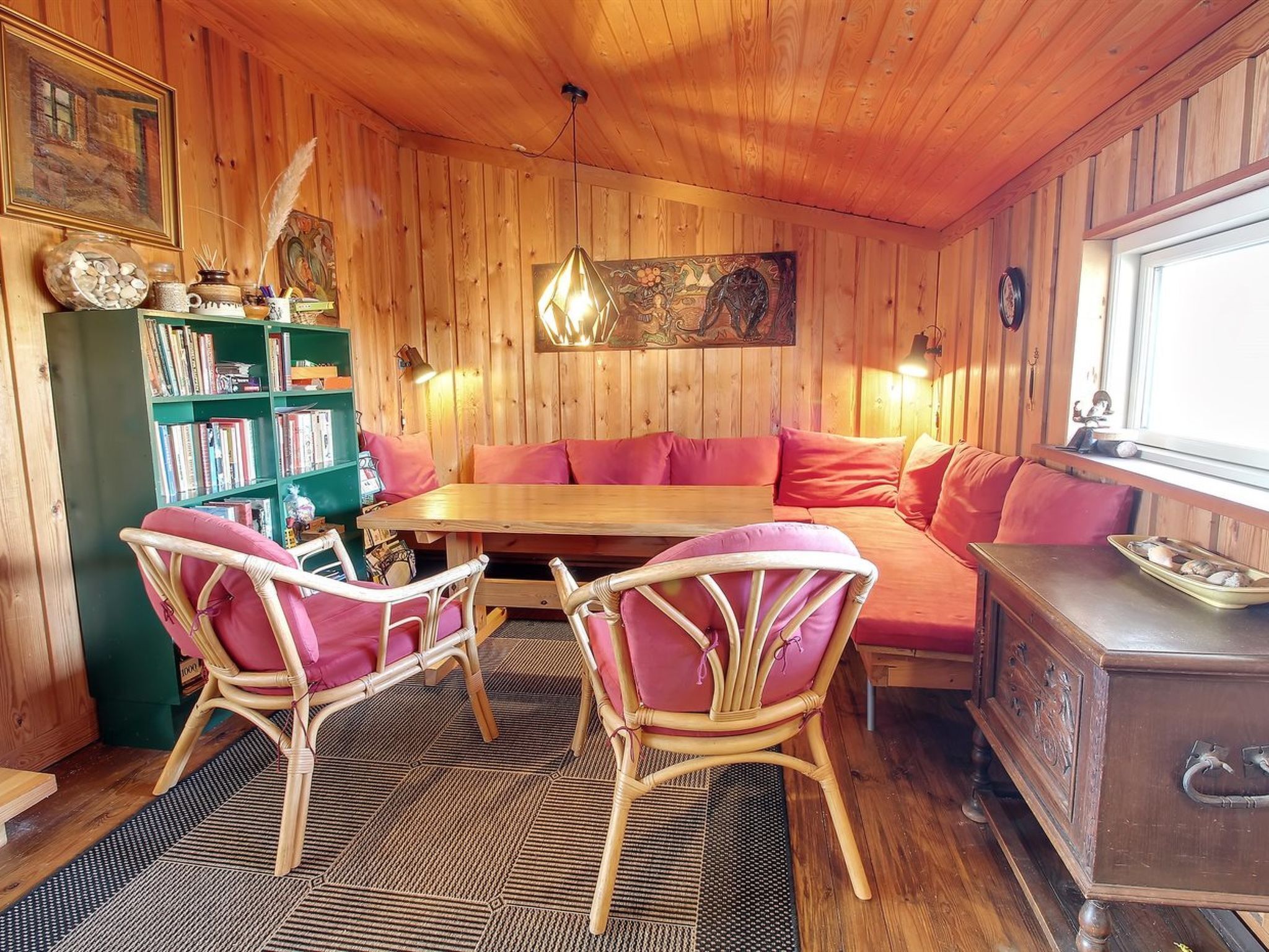 Ein gemütliches Zimmer mit Holzwänden, einem grünen Bücherregal, einem Holztisch, Korbstühlen, einer Couch und einem Holzkasten.