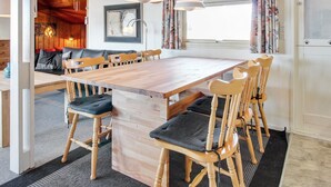 A spacious dining area with a wooden table and chairs, a sofa in the background, and a large window with curtains.