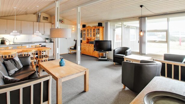 A modern interior with a wooden ceiling, a dining area with a table and chairs, a living room with a sofa and a TV, and a kitchen with cabinets and a stove.