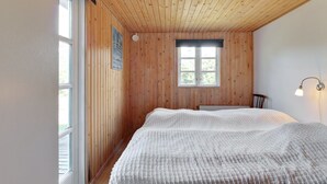 Une chambre avec des murs en bois, une grande fenêtre, un lit recouvert d’une couverture blanche et une lampe fixée au mur.
