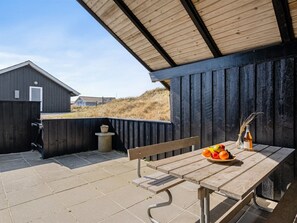 Een terras buiten met een houten tafel, een bankje en een uitzicht op een gebouw en duinen op de achtergrond.