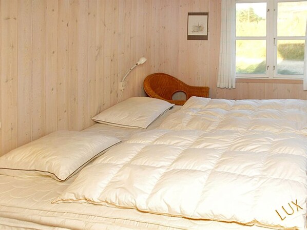 Un lit avec une couette et des oreillers blancs, une tête de lit en bois, une lampe de chevet et une fenêtre donnant sur un paysage verdoyant.