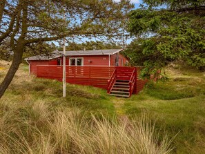 A red cabin with a staircase leading up to a door, surrounded by grass and trees.