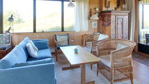 Una habitación acogedora con techo de madera, un sofá con cojines azules, sillas de mimbre, una mesa y un aparador con adornos.