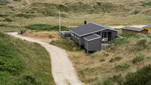 A modern house with a dark roof, situated on a dirt path surrounded by grassy dunes.
