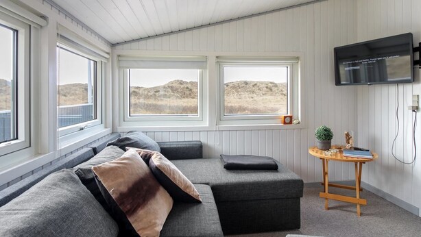 A modern living room with a grey sectional sofa, two windows with views of a dry landscape, a wooden side table with a plant, and a mounted TV.