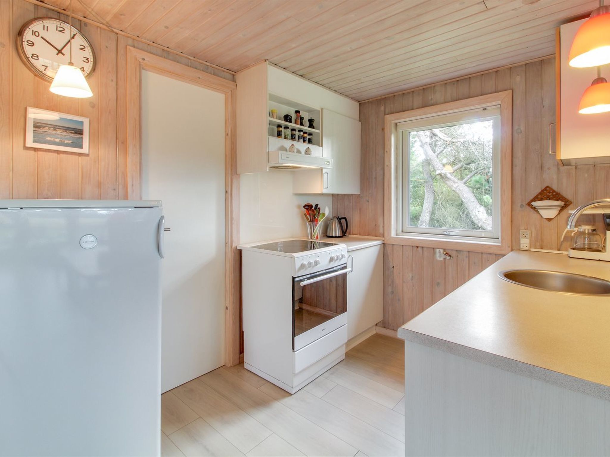 Eine kleine Küche mit Kühlschrank, Backofen und Spüle, Holzwänden und einem Fenster mit Blick auf Bäume.