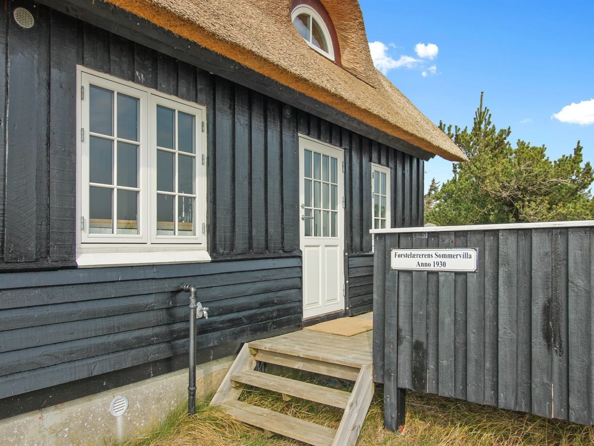 Ein hölzernes Gebäude mit einem Strohdach, zwei Fenstern mit weißen Rahmen und einer Holztür. Ein Schild weist darauf hin, dass es sich um eine Forstlagerersommervilla aus dem Jahr 1930 handelt.