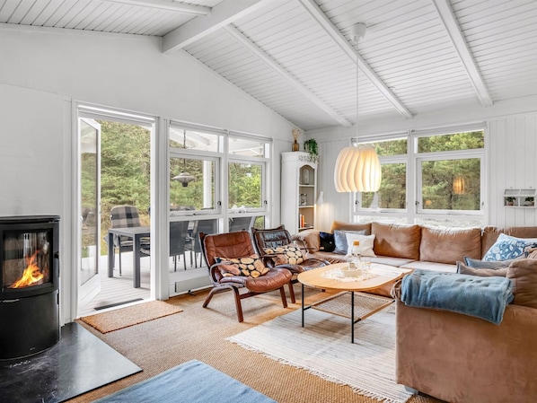 A cozy living room with a fireplace, leather chairs, a sofa, and large windows offering a view of greenery.