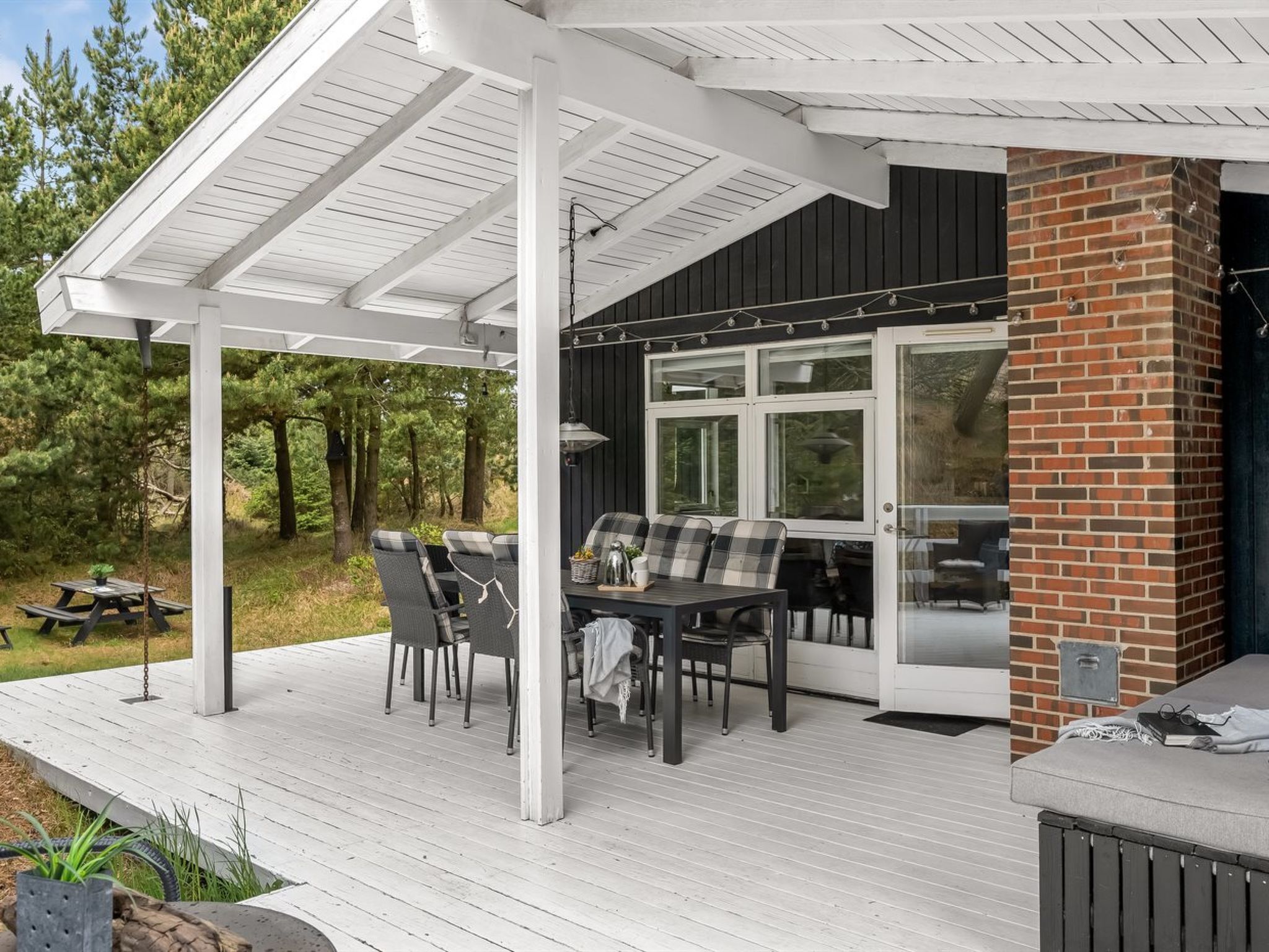 A covered outdoor patio with a table and chairs, a brick fireplace, and a view of the surrounding forest.