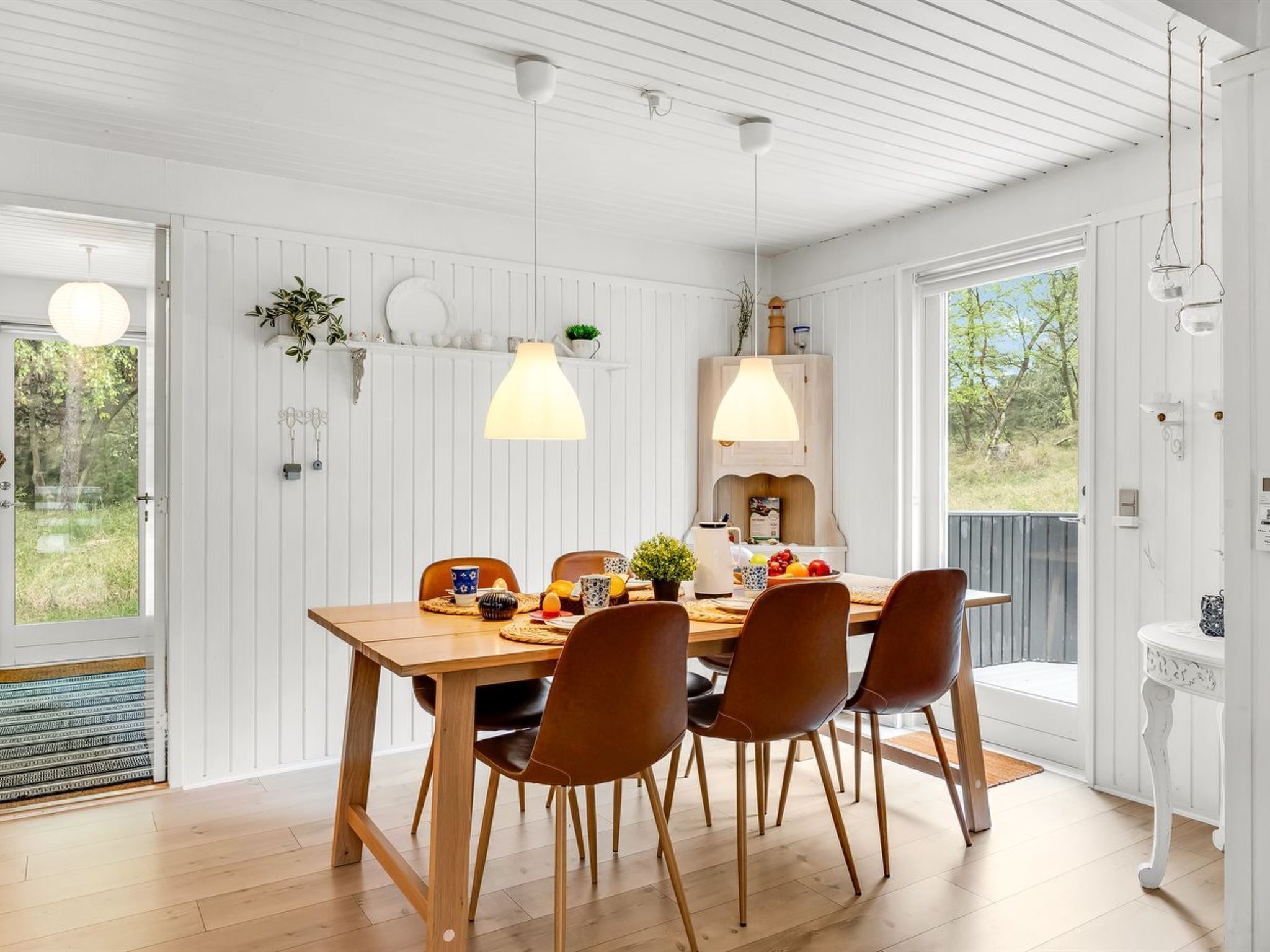Un espace repas comprenant une table en bois, quatre chaises, des luminaires suspendus et une vue sur l’extérieur.