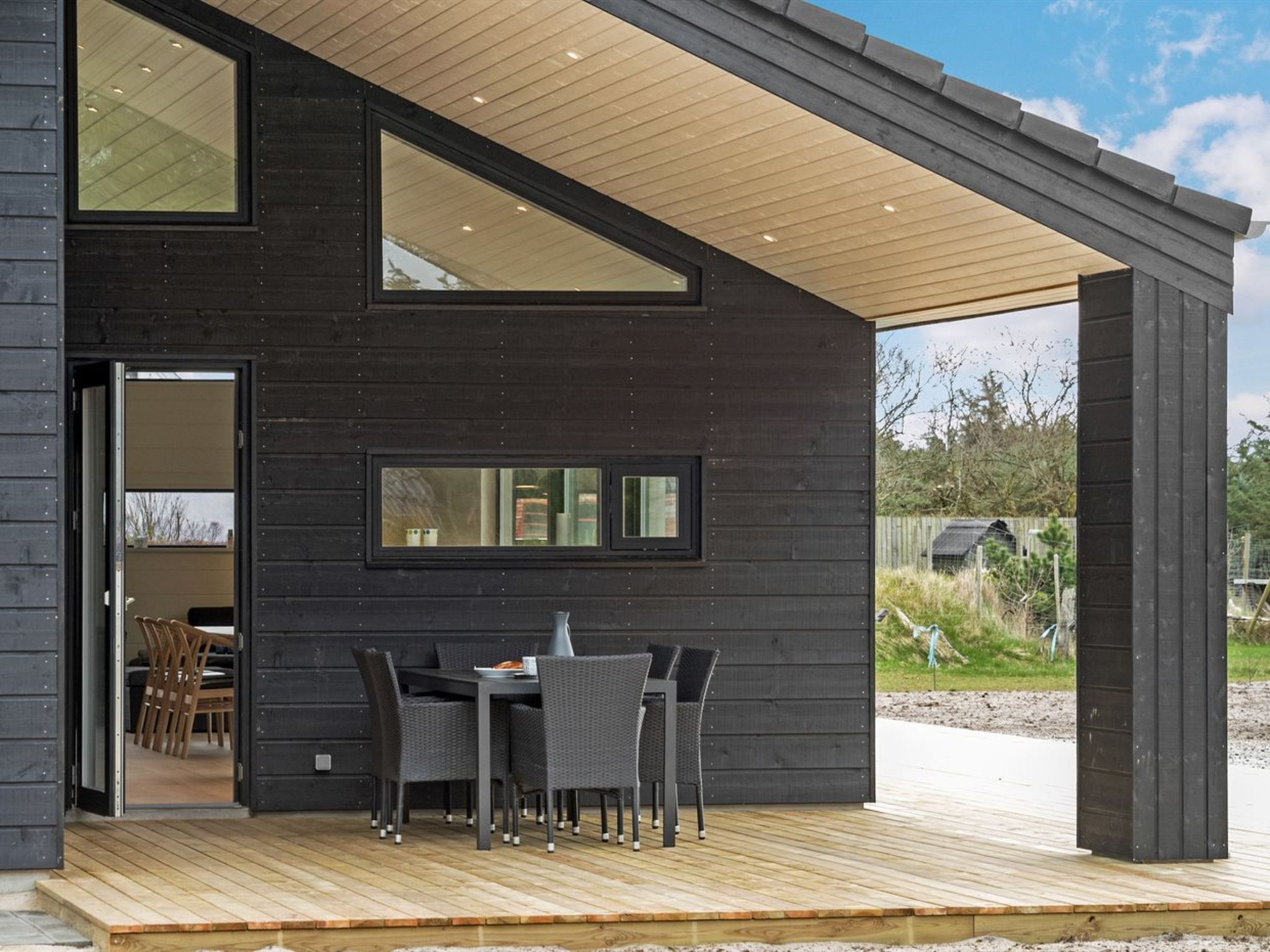 Una casa moderna con terraza de madera, un comedor con sillas y una mesa, y un ventanal grande.