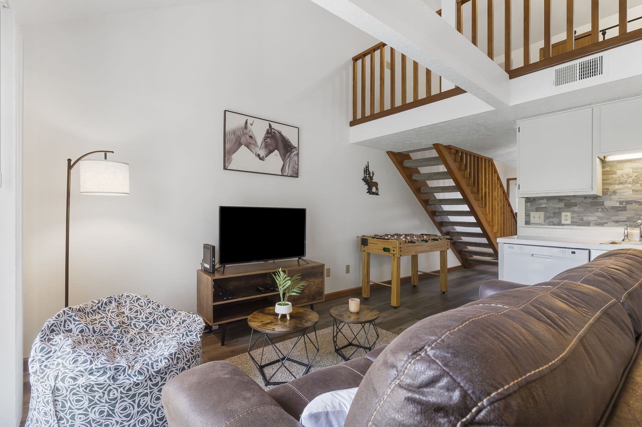 A modern living room with a sofa, a television, a foosball table, and a staircase leading to an upper level.