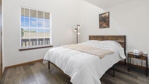 Un dormitorio con una cama grande, una ventana con persianas, una mesita de noche con una lámpara y un cuadro en la pared.